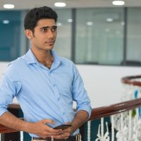 Serious pensive young man with smartphone leaning on railing in lobby and looking into distance. Handsome Indian businessman deep in thoughts using gadget. Planning concept
